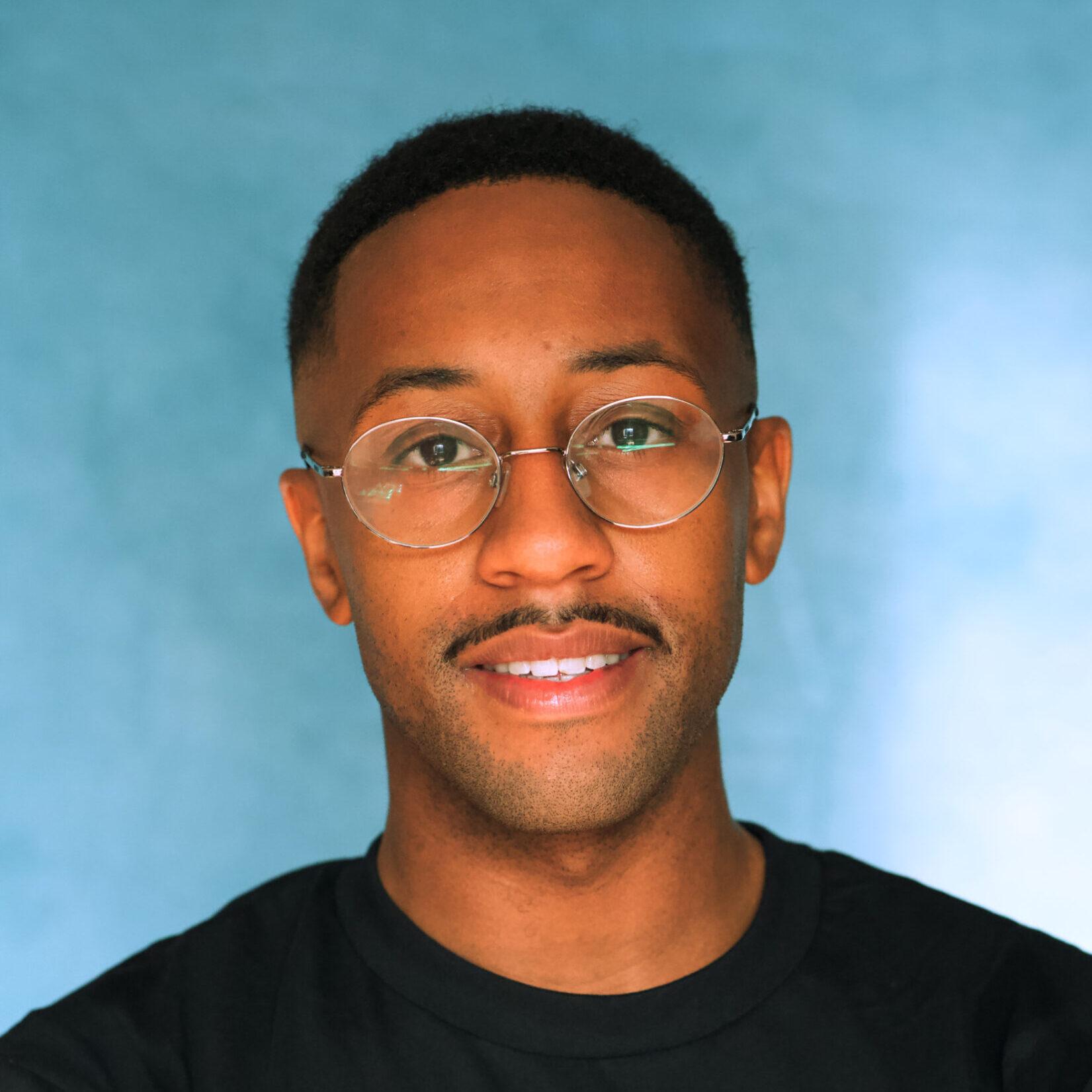Tashan Nicholas Tashan is a Speaker, coach and workshop facilitator at The Hobbs Consultancy who specializes in empowering underserved leaders to bring their authentic selves to the forefront. He is looking at the camera and wears a black top and metal framed glasses.